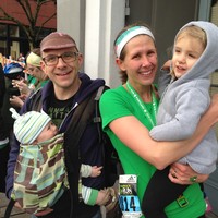 Casual portrait of a young family taken just after the finish at a marathon. The man wears a baby carrier with a young infant. The woman is wearing green running gear and a marathon bib; she holds a preschool age child