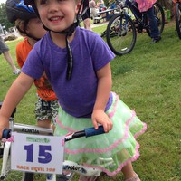 Preschooler wearing a frilly skirt and a bike helmet with a “scoot” pedal-less bicycle. Bike has a racing number that reads “Portland Short Track #15 - Race Devil !@#”