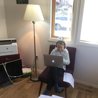A school age child with a laptop and headphones sitting in a chair near a window and a laser printer