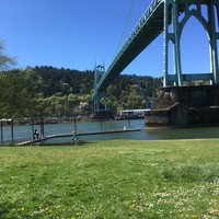 Landscape of Cathedral park from the underside of the St. Johns bridge, looking south/west toward Forest Park. In the midground two children ride bicycles across a broad lawn covered with English lawn daisies. Sky is clear; river is cool and green; some industry visible on the far shore