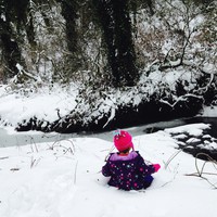 Preschool child in colorful snow gear sitting in the snow next to a partially-icy stream in the forest