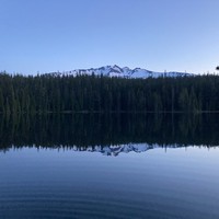 Landscape across a still forest lake at twilight. A single ring ripples slowly across the lake. On the horizon, looming above the trees on the far shore, is the glaciated peak of a broad razorback mountain. Excepting the ripples the lake presents an almost perfect mirror of the scene