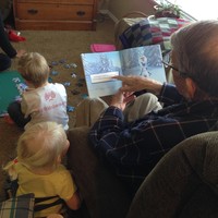 View over the shoulder of an old man reading a FROZEN picture book to a very young toddler. A woman and a young school age boy are putting together a FROZEN puzzle on the floor