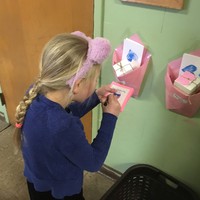 Kindergartener with braids and fuzzy animal ear headband hastily scribbling on Valentine’s day cards outside a classroom