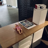 A mostly-bare desk in a darkened, empty office. 2 notable groups of objects on this desk: 1. a plastic baby doll marked “TolaData,” 2. a small pile of personal photographs & effects