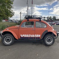 Driver’s side a VW Beetle, Baja mod, with “Speedvagen” painted on the side, and a roof rack. Parked near to the registration tents at a bicycle race