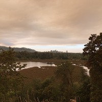 Landscape taken from the top of a bluff across a swampy lake (Oaks Bottom), with the skyline of downtown Portland on the horizon. The air at eye level is crystal clear but overhead at high altitude an unnatural orange cloud of wildfire smoke drifts north toward downtown. The light is orange and eerie.