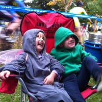 Two young children in sweatshirts (one wearing fairy wings) in a camp chair branded “Laurelwood Brewing Co.” at a bicycle race. They are smiling/laughing at someone off-camera. Very close behind them are two bicycle racers mid-race; to their collective left is a keg of beer in a huge ice bucket