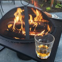 Closeup of a tumbler of whiskey on the margin of a metal above-ground “portable” firepit, with a blazing fire