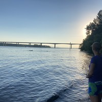 Landscape view westward on the north bank of the Columbia River at sunset. The 205 bridge is on the horizon. On the shore to the right a woman looks out onto the water, where a few hundred feet distant a group of people are swimming