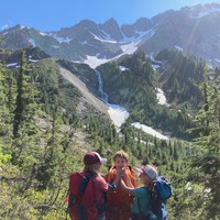 Steep mountain landscape in brilliant sunshine with three kids in backpacks. Two of the kids are using their fingers to force the third’s mouth into a smile shape. About a mile distant are three hanging valleys leading to a lightly glaciated row of peaks; a waterfall drains the topmost glaciers into an ice-covered lake in the lowest valley