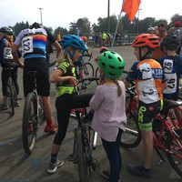 A crowd of children on bicycles, in racing gear, all facing away from the camera, staging for a race. Three kids are the focus of this shot in center frame, in conversation