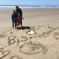 Shot on a beach of a woman and three children in front of a temporary shrine made in the sand. Reads: “Bismarck we love you” with a large heart made from Bismarck’s ashes, and other small bits from the beach like sticks, rocks, and shells