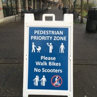 Sandwich board sign on an empty sidewalk, reading “Pedestrian Priority Zone: Please walk bikes / no scooters”